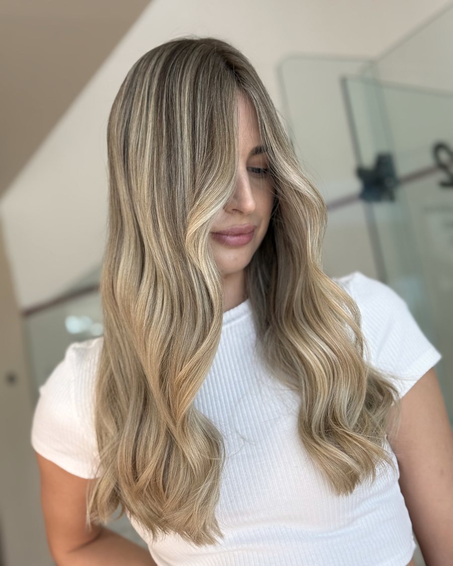 Long hair with a centre parting, blonde balayage from darker roots to lighter ends, styled in soft waves.