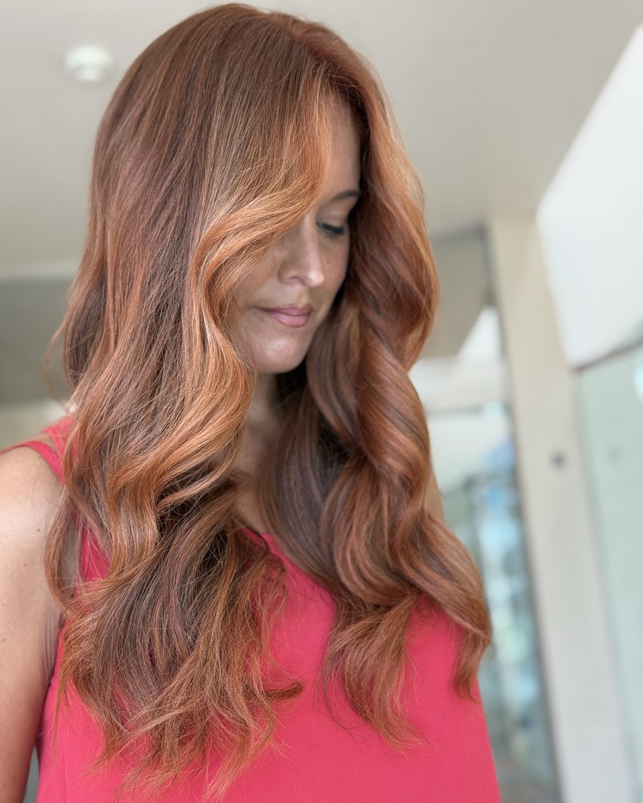 Long hair in a warm copper red with golden highlights, styled in loose waves from a side parting.